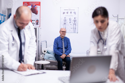 Worried doctors checking health of patient using laptop, anlysing result of patient while senior man waiting in background. Doctor showing and explaining treatment to his colleague using notebook