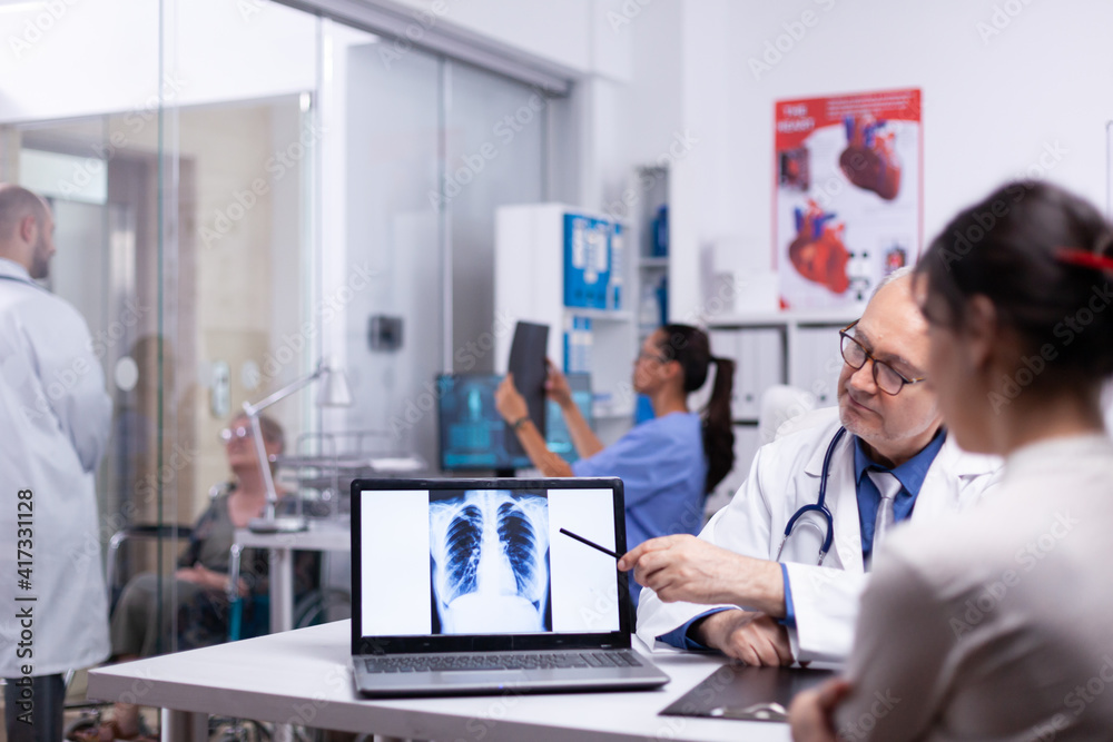 Doctor and nurse analising digital lung x-ray of patient sitting in medical office. Senior physician showing radiography result to young woman pointing with pancil on desktop discussing in front of pc