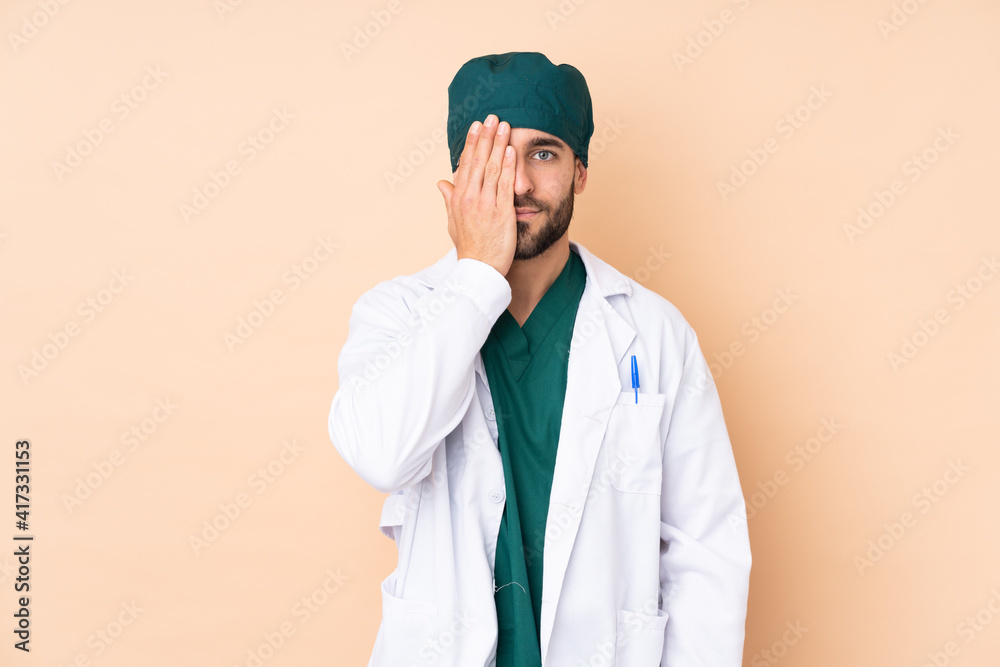 Surgeon man isolated on beige background covering a eye by hand Surgeon man isolated on beige background covering a eye by hand