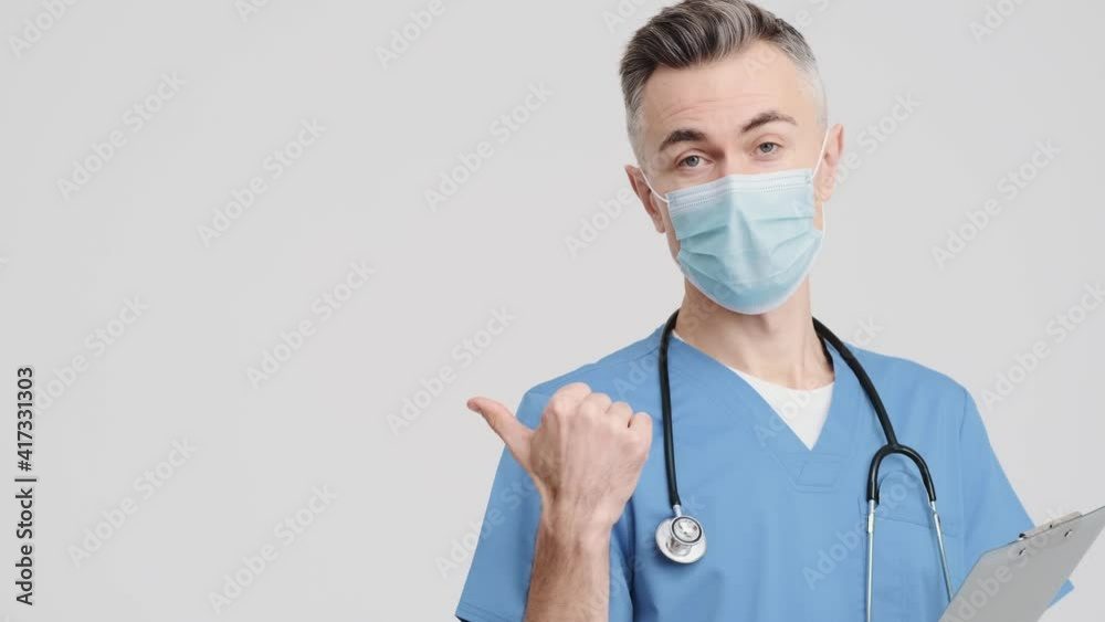 A handsome senior doctor man wearing medical uniform is pointing to the side isolated over grey background in the studio A handsome senior doctor man wearing medical uniform is pointing to the side isolated over grey background in the studio