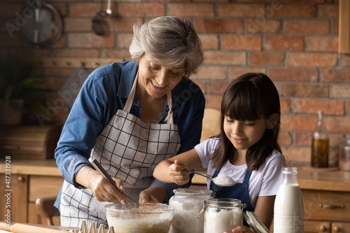 Smiling mature Hispanic granny and small granddaughter work with flour bake cookies in kitchen at home. Happy caring senior grandmother and little grandchild cook breakfast or dessert together.
