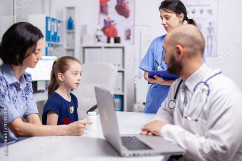 Medical staff talking about treatment with child during expertise in clinic office. Therapeutist in white coat using laptop at consultation for writing medicine prescription