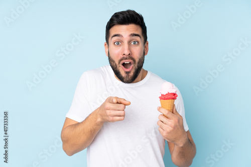 Caucasian man with a cornet ice cream isolated on blue background surprised and pointing front