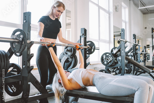 Slim woman in gym with personal trainer exercising power workout with a barbell lying on bench. photo