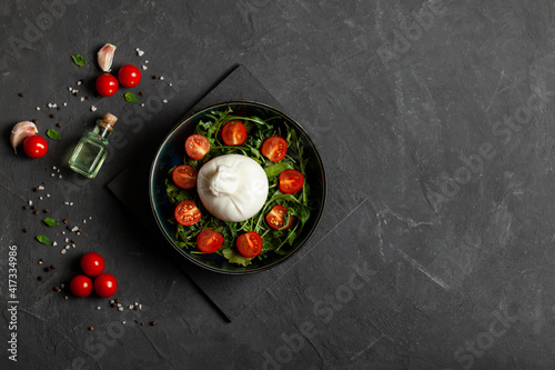 Salad with traditional italian burrata cheese with arugula and tomatoes om dark concrete table with cooking ingredients cherry tomatoes, basil, garlic and olive oil. Top view. Copy space. 