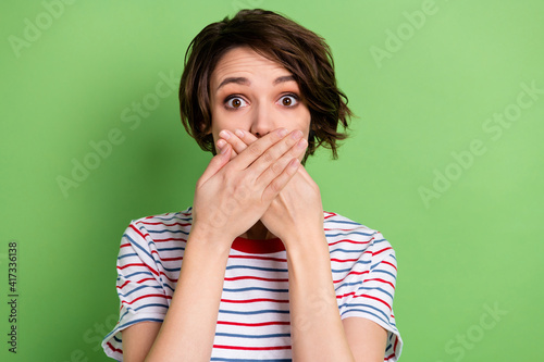 Photo of worried young girl arms palms covering mouth lips staring camera isolated on green color background