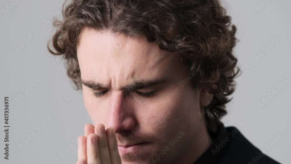 A nervous young man is holding hands on a praying gesture standing isolated over gray wall in the studio A nervous young man is holding hands on a praying gesture standing isolated over gray wall in the studio