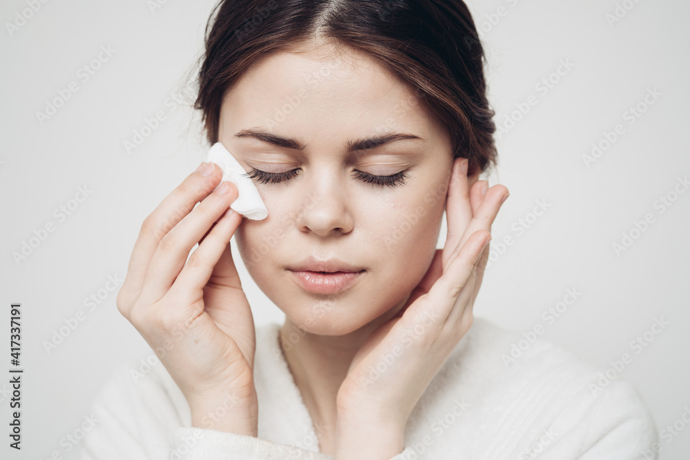brunette woman wipes her face with a white sponge on a light background brunette woman wipes her face with a white sponge on a light background