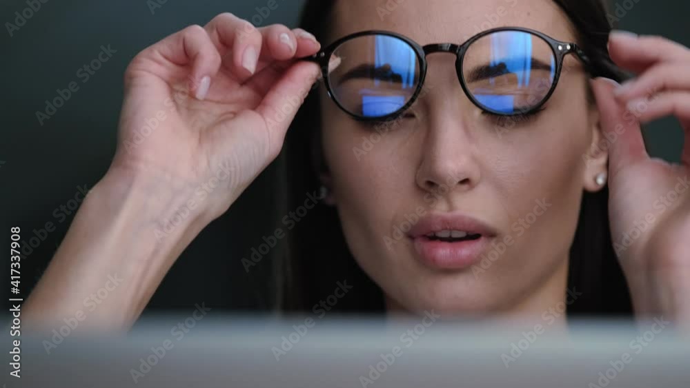 A close-up view of a concentrated young woman is using her laptop while putting off her eyeglasses sitting at the apartments A close-up view of a concentrated young woman is using her laptop while putting off her eyeglasses sitting at the apartments