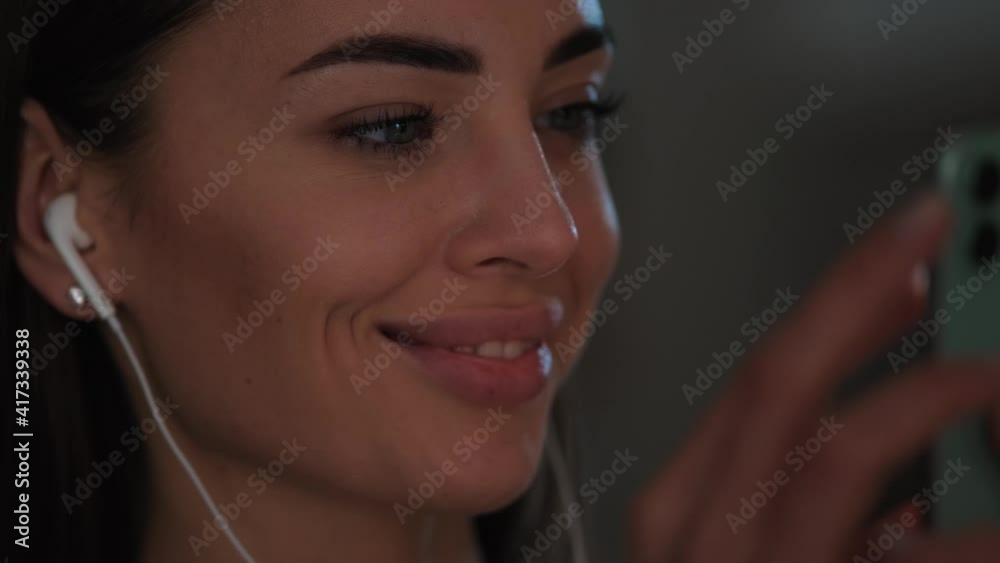 A close-up view of a smiling woman wearing earphones is watching something in her smartphone inside the apartments A close-up view of a smiling woman wearing earphones is watching something in her smartphone inside the apartments