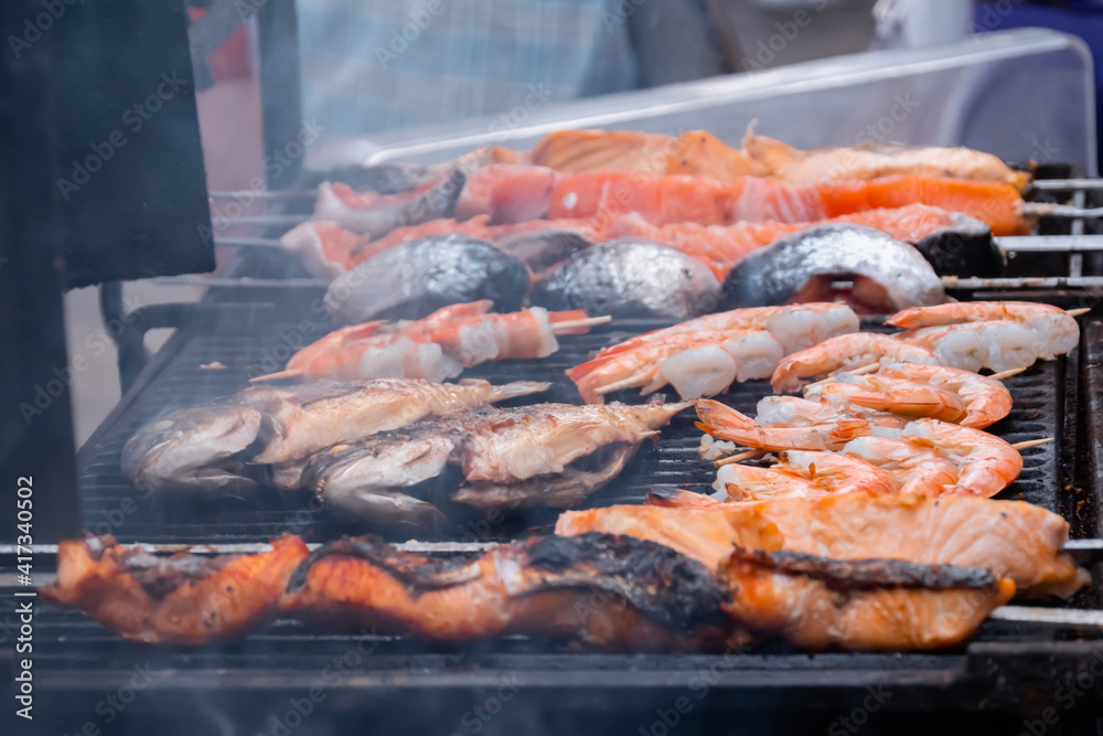 Cooking salmon, tiger shrimp, prawn skewers and mackerel fish on grill at summer local food market - close up. Outdoor cooking, barbecue, gastronomy, seafood, cookery, street food concept