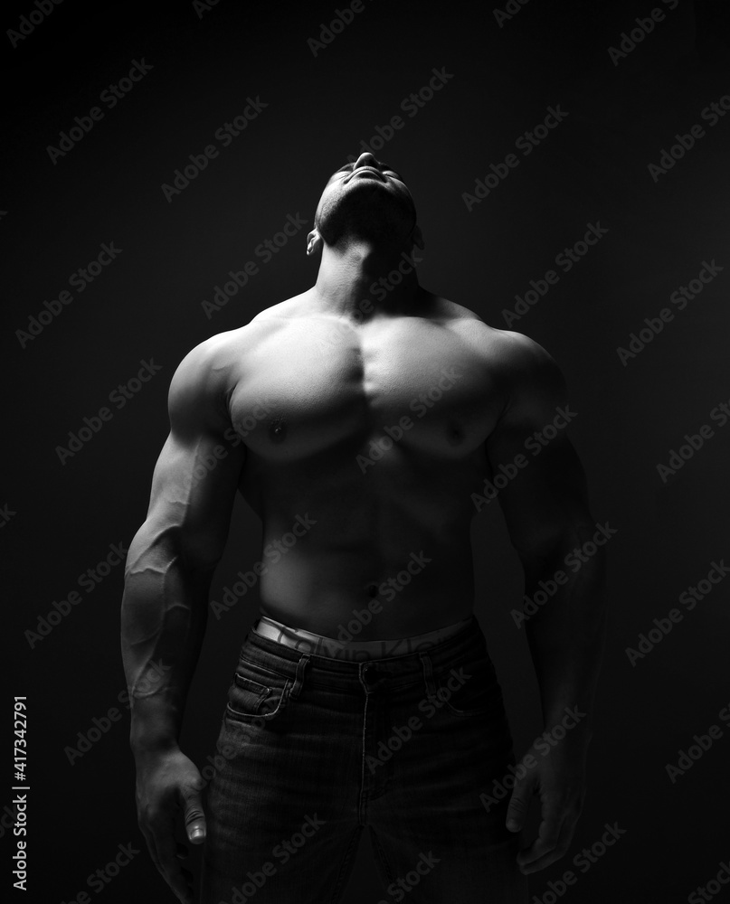 Black and white portrait of handsome muscular man, athlete with perfect built body stands in jeans and topless, demonstrating chest muscles, with head thrown back, looking up over dark background