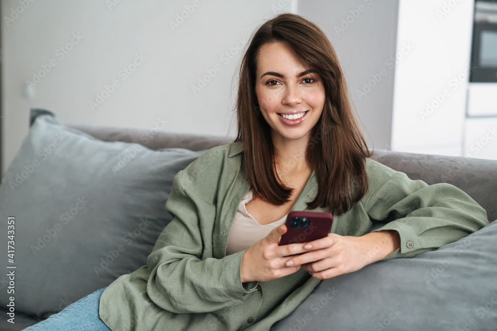 Happy brunette woman smiling and using cellphone while sitting on floor Happy brunette woman smiling and using cellphone while sitting on floor
