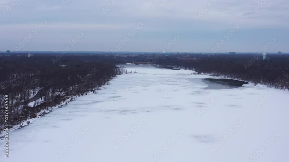 An aerial view from a drone, over a long and wide lake during a cloudy sunrise. The drone camera dolly in over the snow covered, frozen lake below. There are bare tree on either side of the lake. An aerial view from a drone, over a long and wide lake during a cloudy sunrise. The drone camera dolly in over the snow covered, frozen lake below. There are bare tree on either side of the lake.