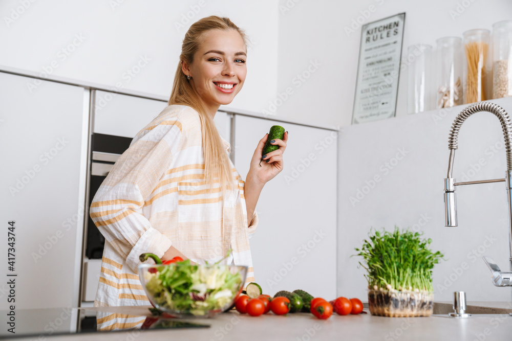 Joyful blonde woman holding cucumber while preparing fresh salad