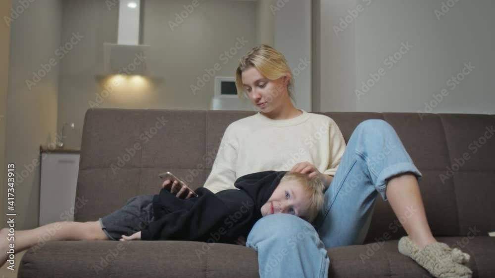 Young mother sits with her son on couch, she uses phone while her son watches TV Young mother sits with her son on couch, she uses phone while her son watches TV