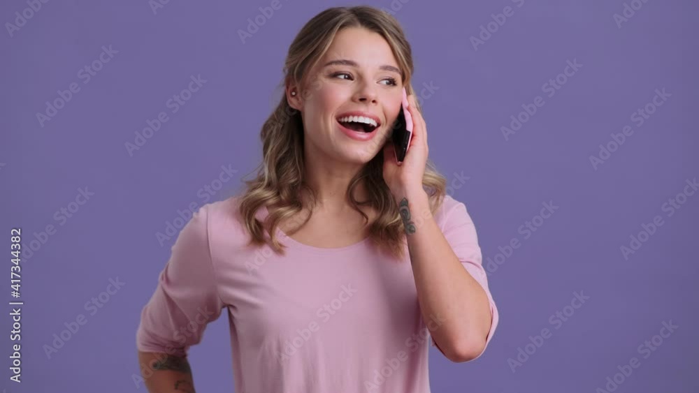 A positive woman is talking on her smartphone standing isolated over a violet background A positive woman is talking on her smartphone standing isolated over a violet background