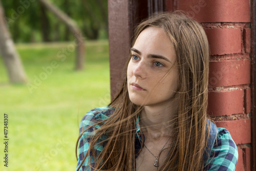 A beautiful young long-haired sad lonely girl in a plaid shirt sits on a park bench in summer or spring.