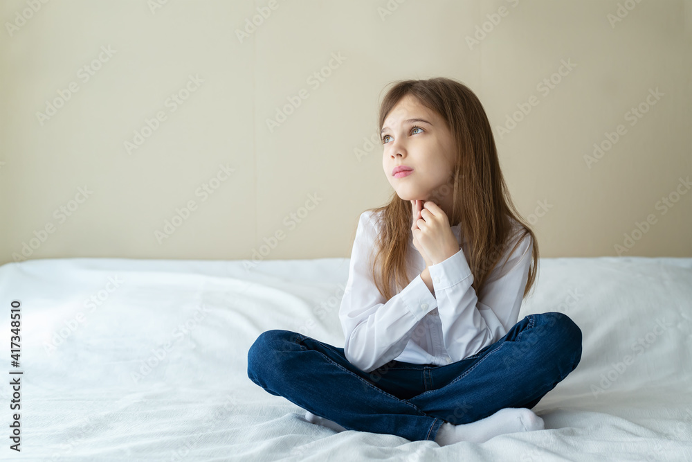 Close up shot of beautiful blonde caucasian little girl wearing jeans and a white shirt, sitting on a bed and looking calm and dreamy Close up shot of beautiful blonde caucasian little girl wearing jeans and a white shirt, sitting on a bed and looking calm and dreamy