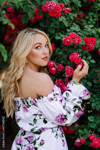 Outdoor photo of romantic young woman in rose garden. girl with red lips in a dress with a print of roses. Young model on a background of a bush of roses. Stylish woman.
