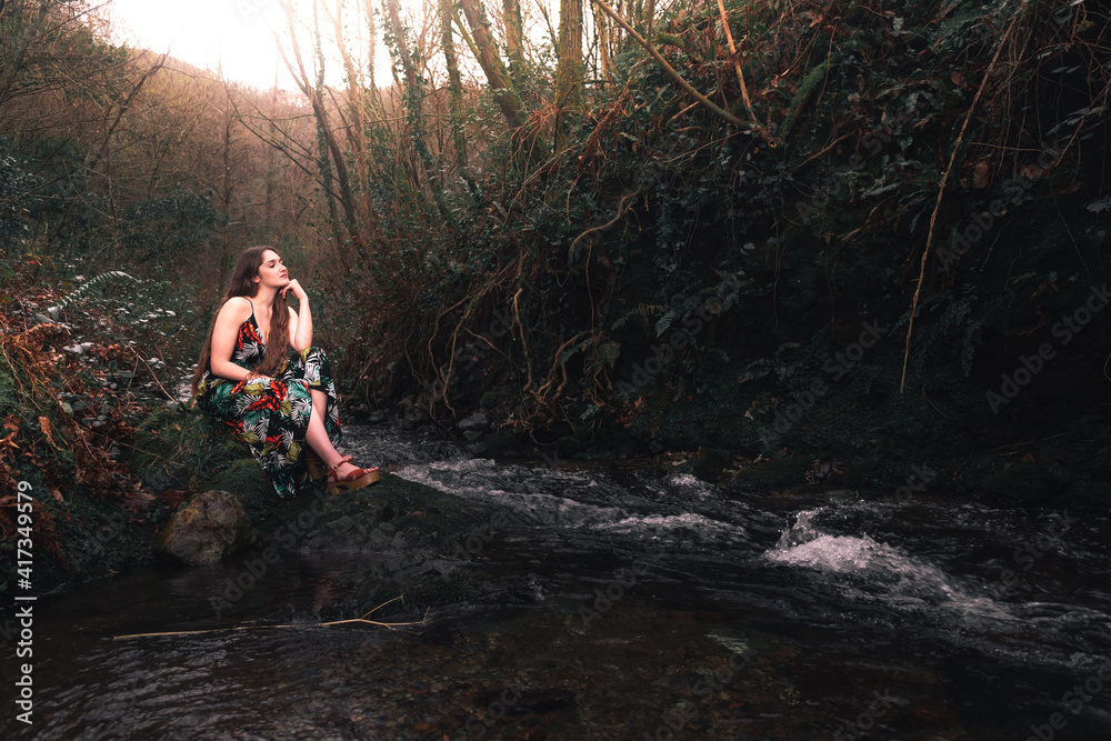 Young caucasian woman with a colorful dress in the middle of the forest next to a little river