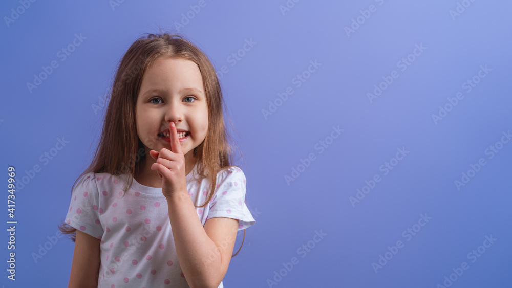 portrait of a cute little girl, holding her index finger to her mouth, stands on a purple background. The child doesn't want to talk. Keeps a secret, learned a secret portrait of a cute little girl, holding her index finger to her mouth, stands on a purple background. The child doesn't want to talk. Keeps a secret, learned a secret