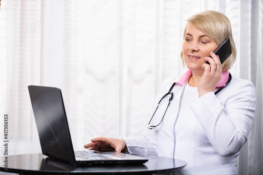 Senior female doctor in medical uniform in front of laptop with phone during online consultation, working day. Senior female doctor in medical uniform in front of laptop with phone during online consultation, working day.