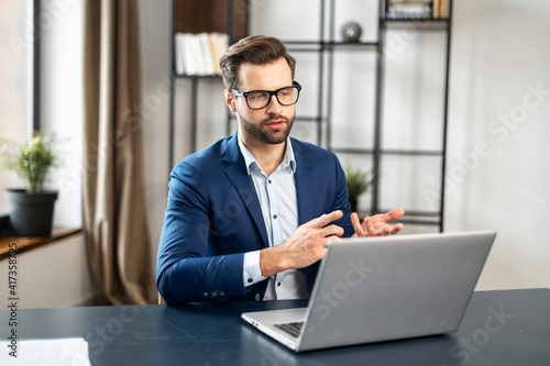 Young bearded entrepreneur, business coach in formal wear having coaching session on leadership and sales or discussing project, gesturing, presenting strategy, idea online on video call via laptop