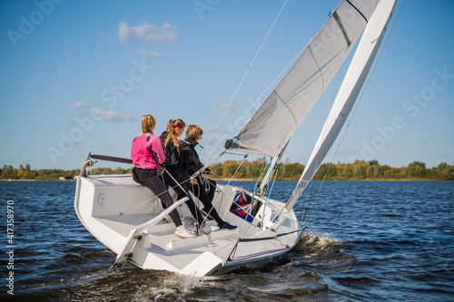 A beautiful white racing single-masted yacht is sailing against a beautiful river landscape with a blue sky. A man and two girls are on board