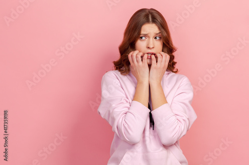 Portrait of worried curly haired teenager girl in hoodie biting nails with panicked terrified face, having depression and problem, anxiety disorder. Indoor studio shot isolated on pink background