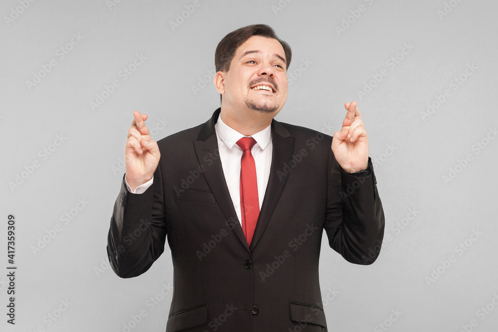 Businessman crosses his fingers and hopes for luck. indoor studio shot. isolated on gray background. handsome businessman with black suit, red tie and mustache looking at camera.