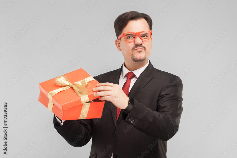 Business man holding red gift box disappointed with the gift. Indoor, studio shot, isolated on gray background Business man holding red gift box disappointed with the gift. Indoor, studio shot, isolated on gray background