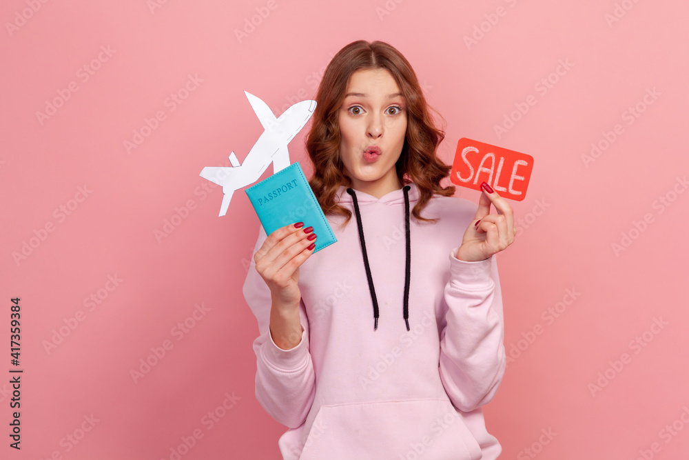 Portrait of excited curly haired teenage girl in hoodie showing passport with paper airplane and sale word sign, buying cheap tickets, discount. Indoor studio shot isolated on pink background Portrait of excited curly haired teenage girl in hoodie showing passport with paper airplane and sale word sign, buying cheap tickets, discount. Indoor studio shot isolated on pink background
