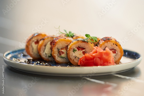 A close-up photo of fresh sushi rolls with salmon, Philadelphia cheese, and tuna on a blue ceramic plate with sauce, sesame, wasabi, and leaves. Black stone table.