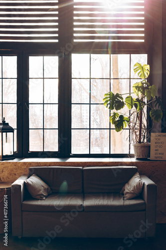 Living room in loft style. Gray sofa on the background of a large window with sunbeams. Spring sun in the room. Modern room interior with dark furniture.