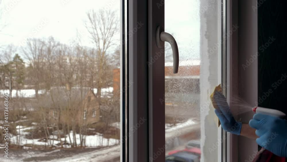 A young guy in a red T-shirt and blue gloves puffs red window liquid, then wipes the windows with a yellow rag. Window cleaner concept