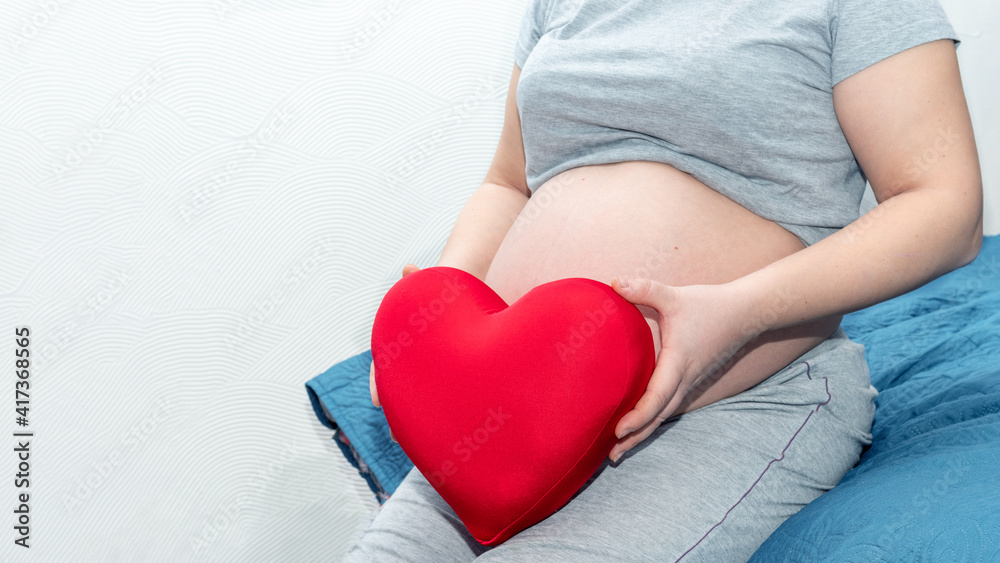 Happy motherhood concept. Female hands hold a red pillow in the shape of a heart against the background of the belly of a pregnant woman. Mothers day concept Happy motherhood concept. Female hands hold a red pillow in the shape of a heart against the background of the belly of a pregnant woman. Mothers day concept