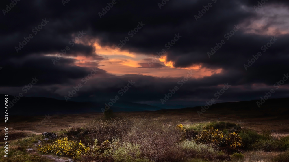 Extreme weather and lighting conditions during sunset in the Scottish Highlands.Dark and dramatic sky with black clouds with a window of orange sunlight over hills.Beautiful landscape of Scotland.
