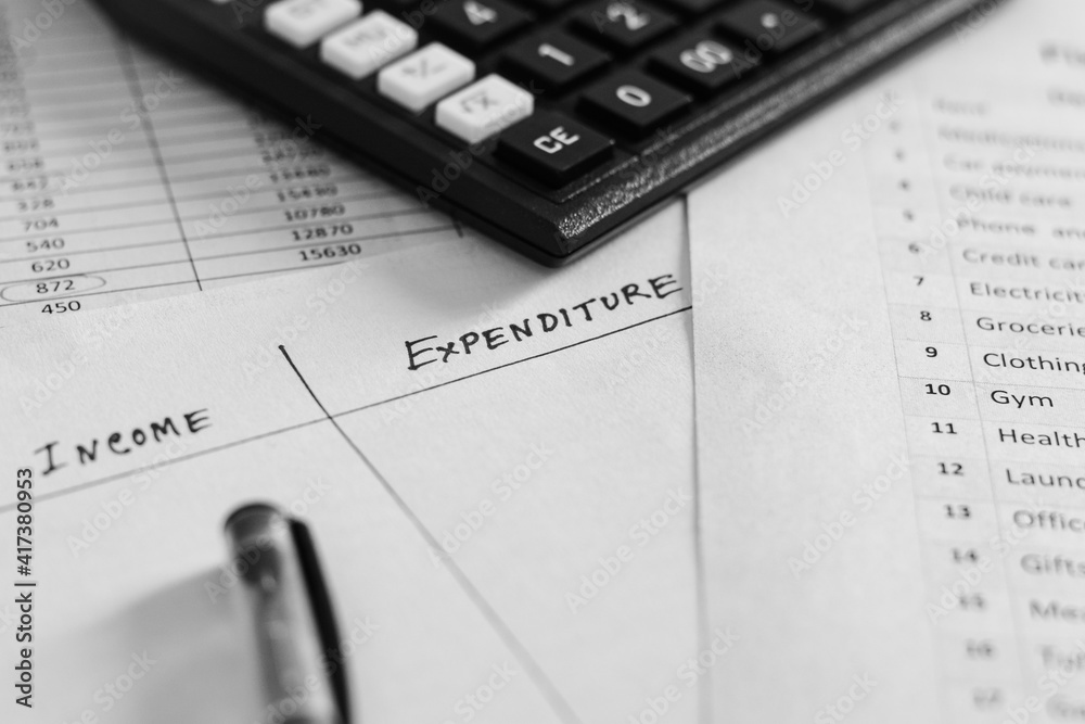 Income and expenditure analysis idea. Handwritten text in notepad placed on page background with expense list, calculator and pen. Selective focus on the text expenditure. Income and expenditure analysis idea. Handwritten text in notepad placed on page background with expense list, calculator and pen. Selective focus on the text expenditure.