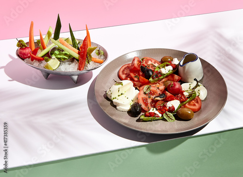 Tomato  olives and mozzarella salad . Restaurant appetizer plate on white table with pink wall. Day sunlight with hard shadow of fern palm leaves. Summer or spring restaurant food concept