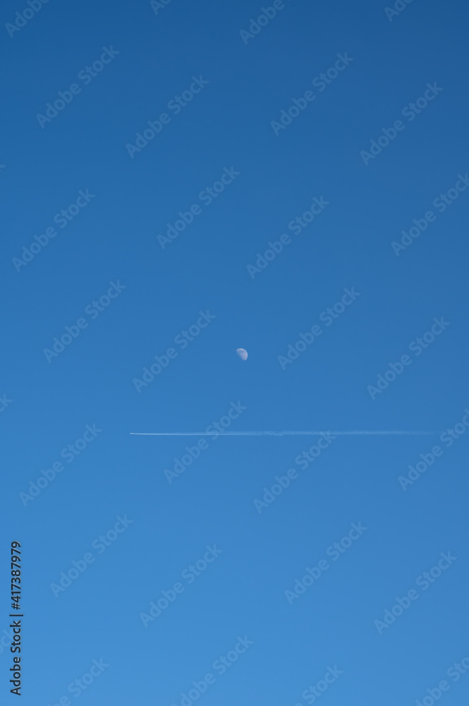 A trail after a plane is seen just under the moon on a clear blue sky A trail after a plane is seen just under the moon on a clear blue sky