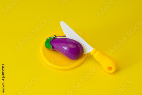 Creative composition of toy eggplant with a knife on a yellow background.