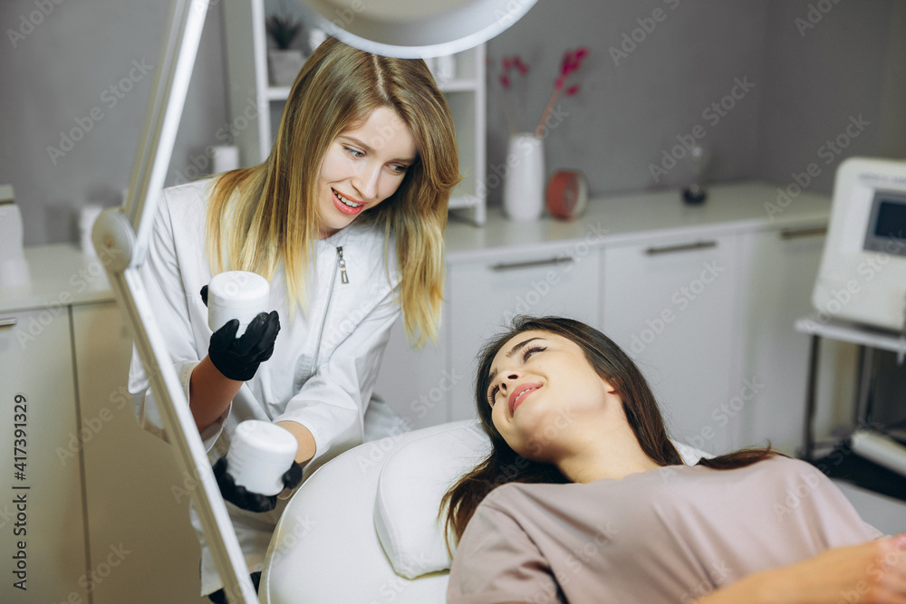 Consultation in cosmetology clinic. Female beauty doctor talking with patient and showing her what she's going to use, cream or mask. Consultation in cosmetology clinic. Female beauty doctor talking with patient and showing her what she's going to use, cream or mask.