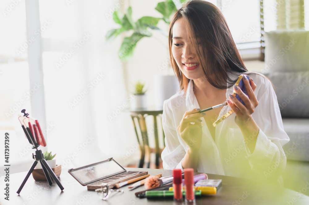 Beauty blogger influencer Asian woman using smartphone live streaming review cosmetics products in living room at home Beauty blogger influencer Asian woman using smartphone live streaming review cosmetics products in living room at home