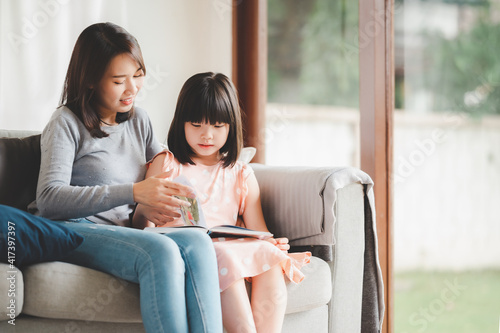 Happy Asian family mother and daughter sitting on sofa reading a book in living room at home