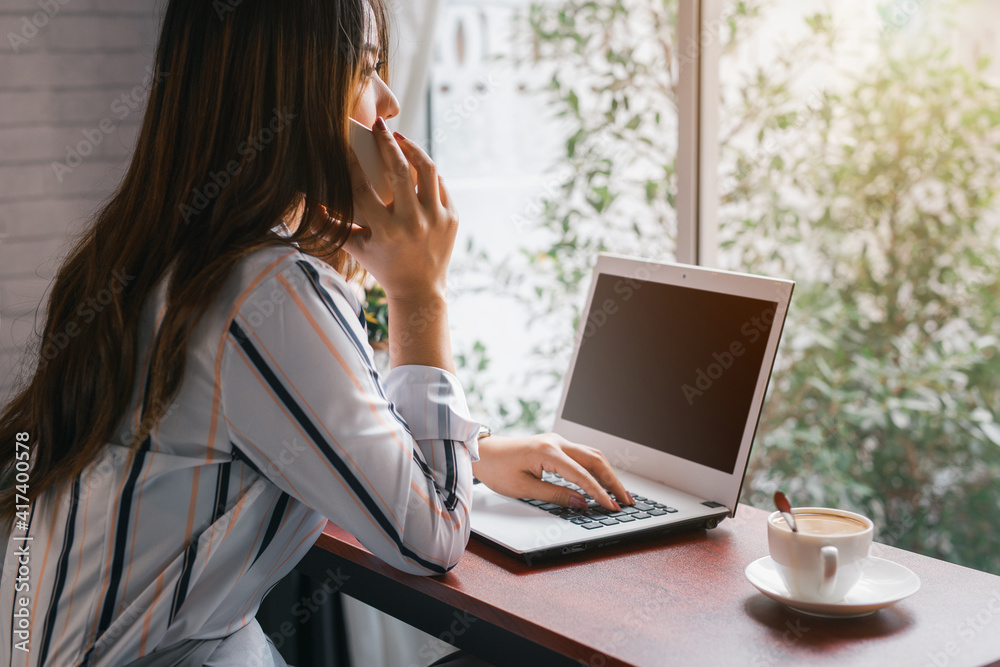 Beautiful young business woman using smart phone and working on laptop computer while sitting drinking coffee at home. Beautiful young business woman using smart phone and working on laptop computer while sitting drinking coffee at home.