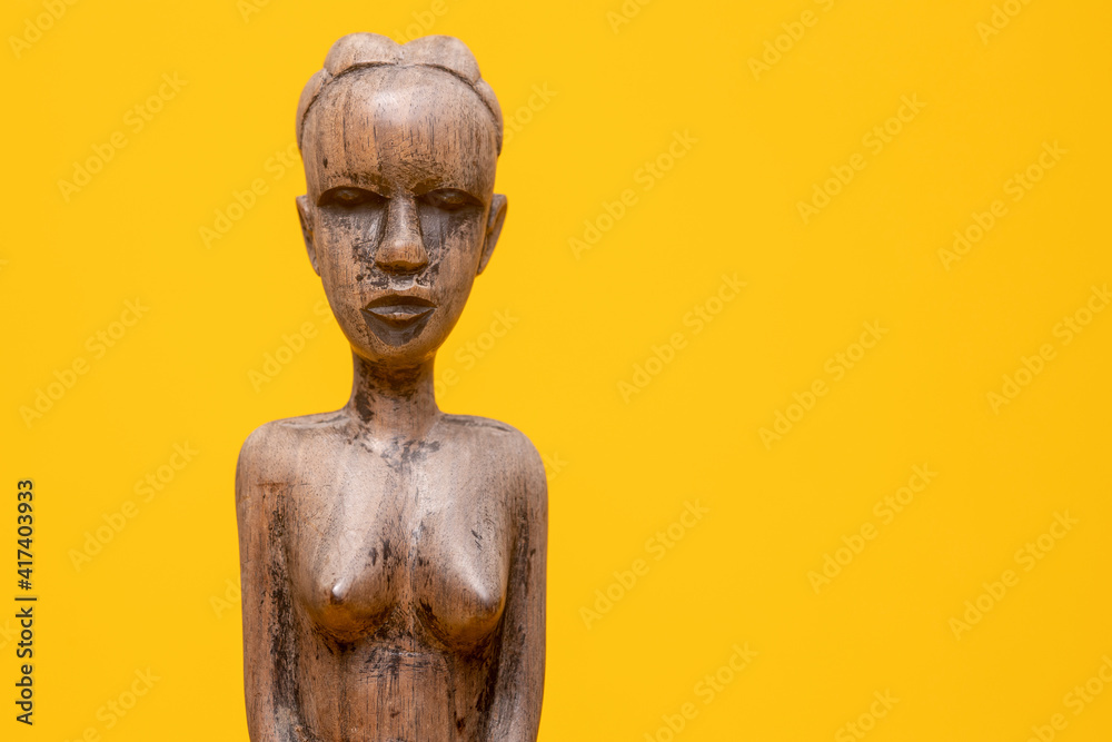 Closeup of head and torso of carved out female figurine of ebony wood without clothes on holding hands in front of her private area against a seamless yellow background.