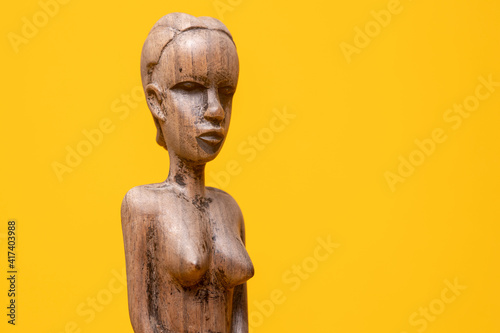 Sideview closeup of female figurine carved out of ebony wood without clothes on holding hands in front of her private area against a seamless yellow background.