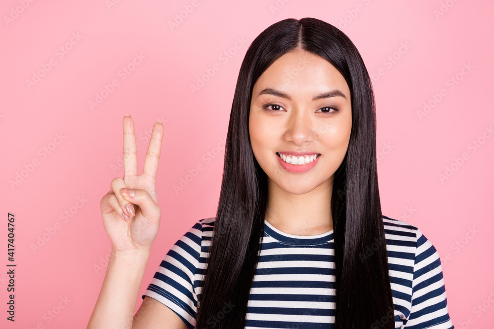 Photo portrait of woman showing v-sign gesture wearing striped t-shirt smiling isolated on pastel pink color background Photo portrait of woman showing v-sign gesture wearing striped t-shirt smiling isolated on pastel pink color background