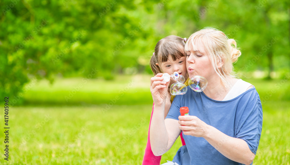 Little girl with syndrome down and her mother blow bubbles in a summer park. Empty space for text Little girl with syndrome down and her mother blow bubbles in a summer park. Empty space for text